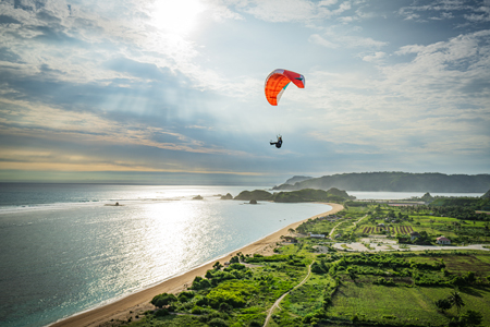 Stunning view of a paraglider flying over the breathtaking landscape of Indonesia, with mountains and coastline in the background, captured in Bukit Waung. The pilot is flying a NOVA Performance Paragliders Doubleskin 2 wing, enjoying a smooth and thrilling flight over a paradise-like setting.